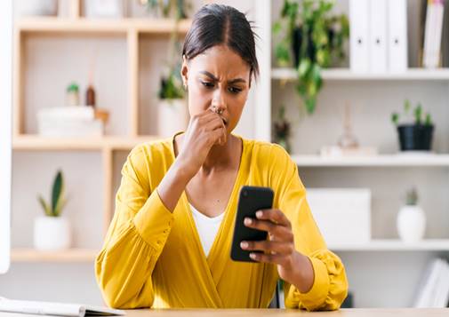 A woman wearing a yellow shirt looks worried while looking at her phone. She is seated at a desk in front of a shelf with books and plants.